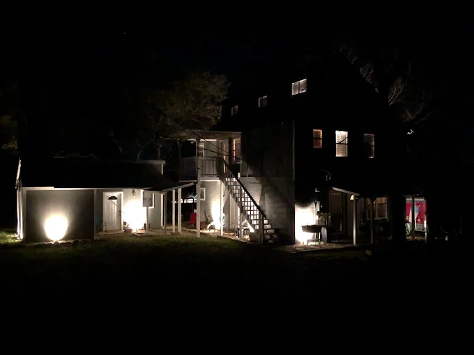 A house is illuminated at night with exterior lights highlighting stairs, windows, and doorways while the surrounding area remains dark.